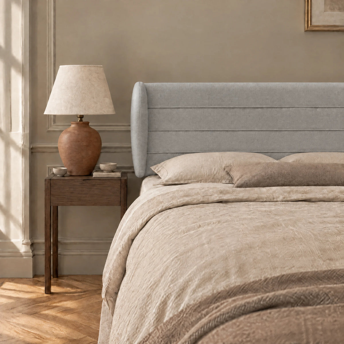 Tête de lit à oreilles en tissu Bagatelle gris clair dans une chambre, avec table de chevet et lampe